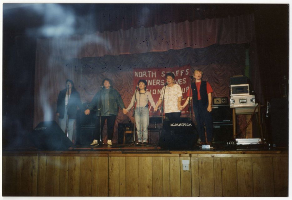 Colour photograph of five women standing on a wooden stage. Behind them hangs a red banner with white text reading “North Staffs Miners’ Wives Action Group.” The women, including Rose Hunter, Brenda Procter, and Bridget Bell, are standing in a line, holding hands in a gesture of solidarity.